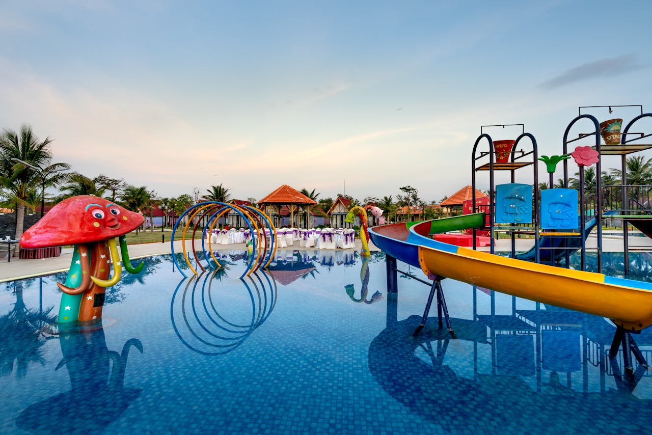 Colorful waterpark with slides, mushroom-shaped feature, and palm trees reflecting in pool.